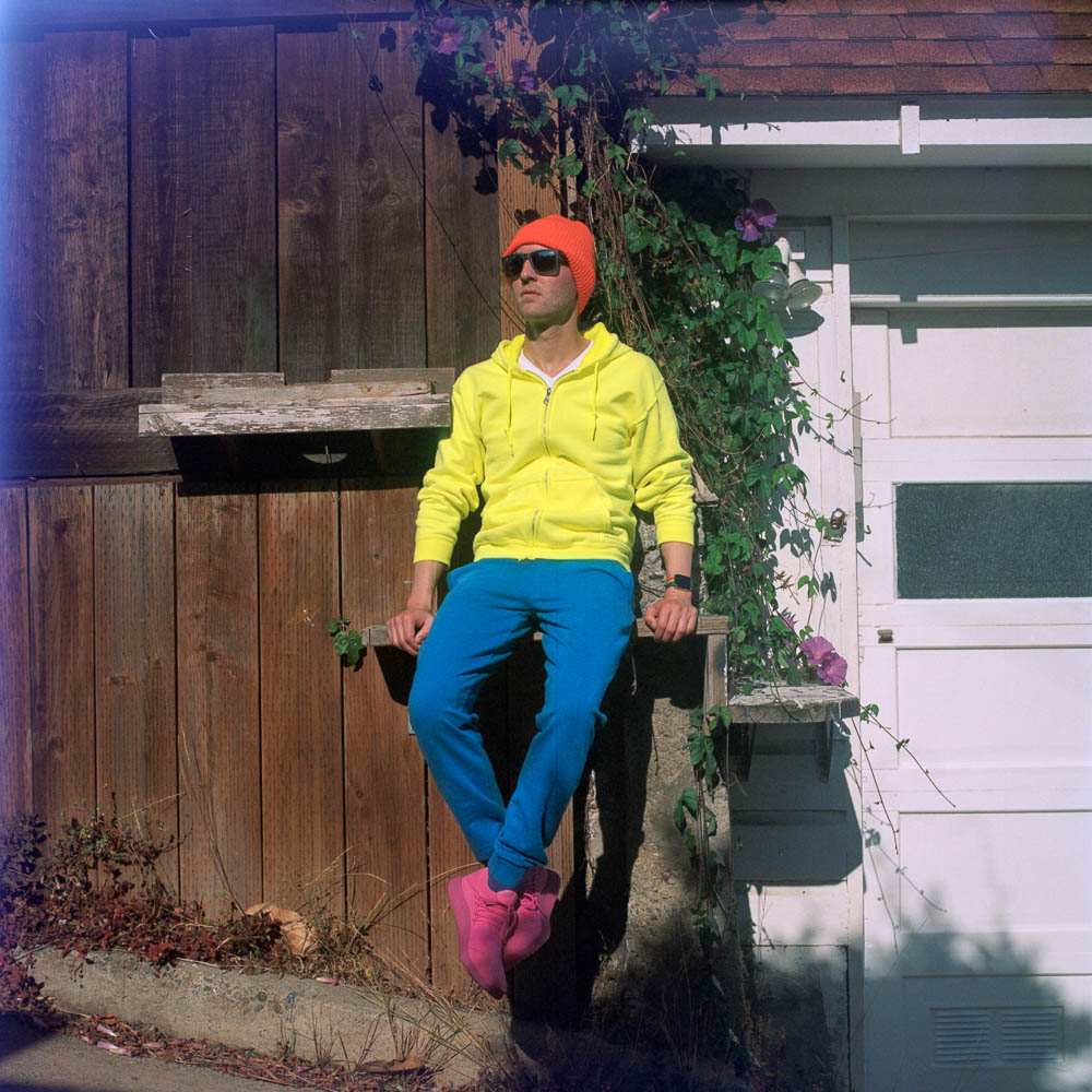 portrait of Marc sitting on a wooden post against a wooden fence, wearing neon